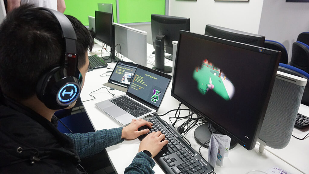 A photo of someone wearing headphones as they play Honeycomb Festival on the desktop and keyboard. On the screen is a copy of the game, showing Lynette, a pink fairy, wandering across a green field. There is a blindness filter overlaid on the game, blocking out the edges of the screen in black, to simulate Glaucoma.