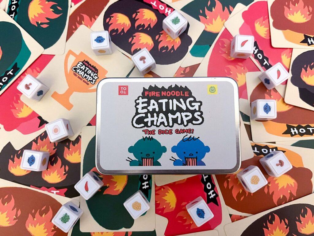 A photo of the Fire Noodle Eating Champs dice game container, sat on the various cards and die from the game. 