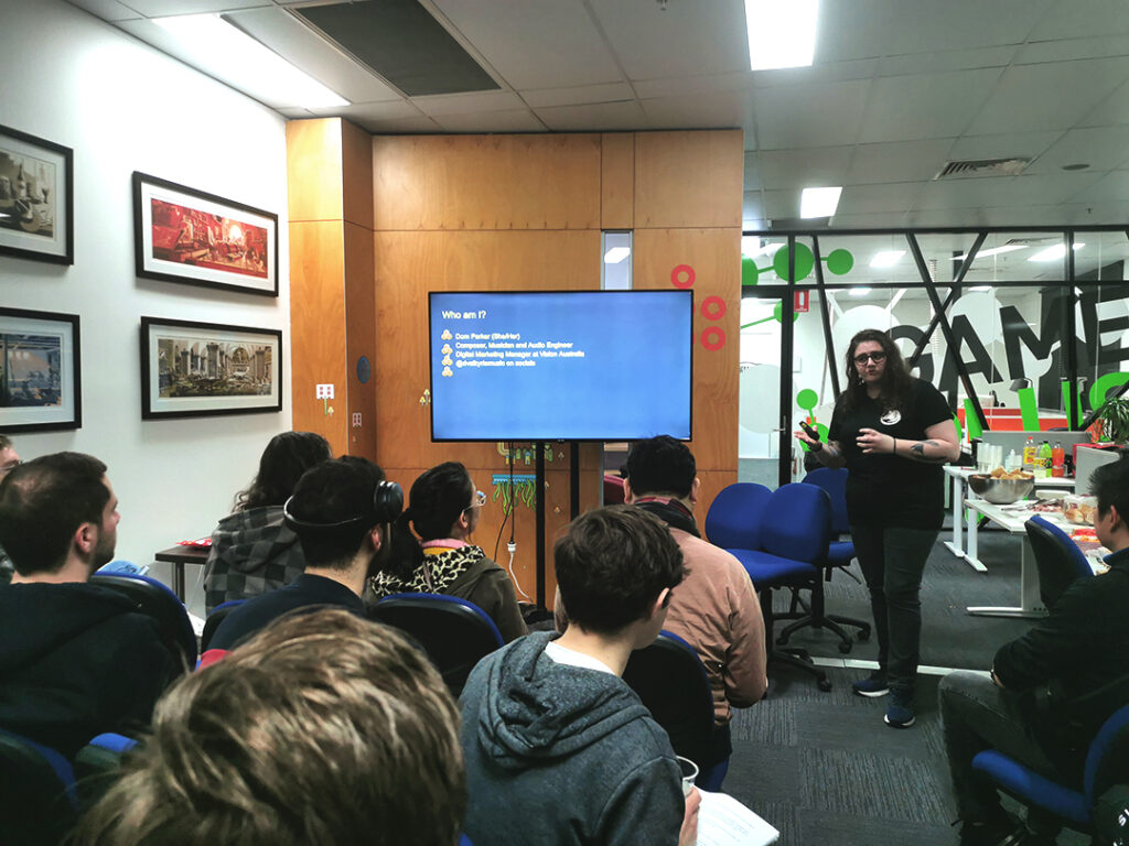 A photo of Dom presenting in front of a crowd at Game Plus. Next to her is a large tv screen showing off a slide about her personal details: Such as her name and pronouns, position (Composer, Musician and Audio Engineer, Digital Marketing Manager at Vision Australia) and her socials (@DValkyrieMusic).