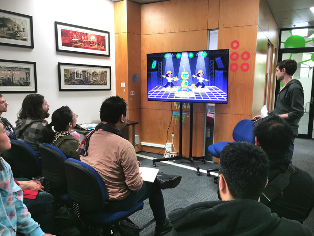 A photo of Evan presenting the Kickstarter Bits & Bops trailer at WIP Night. On the large tv screen is a shot of some robots and dancers dancing on a dark and neon dance floor.