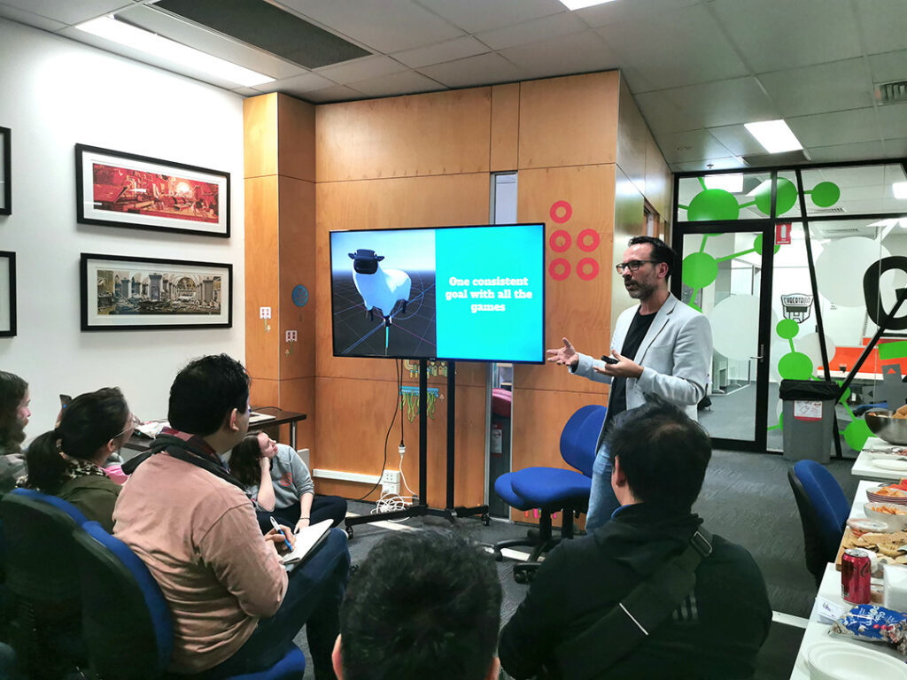 A photo of David presenting about his goals with every Sheep game over the years. On the large tv screen is a blue presentation slide with a screenshot of the sheep in game. Next to it is text that says "One consistent goal with all the games".