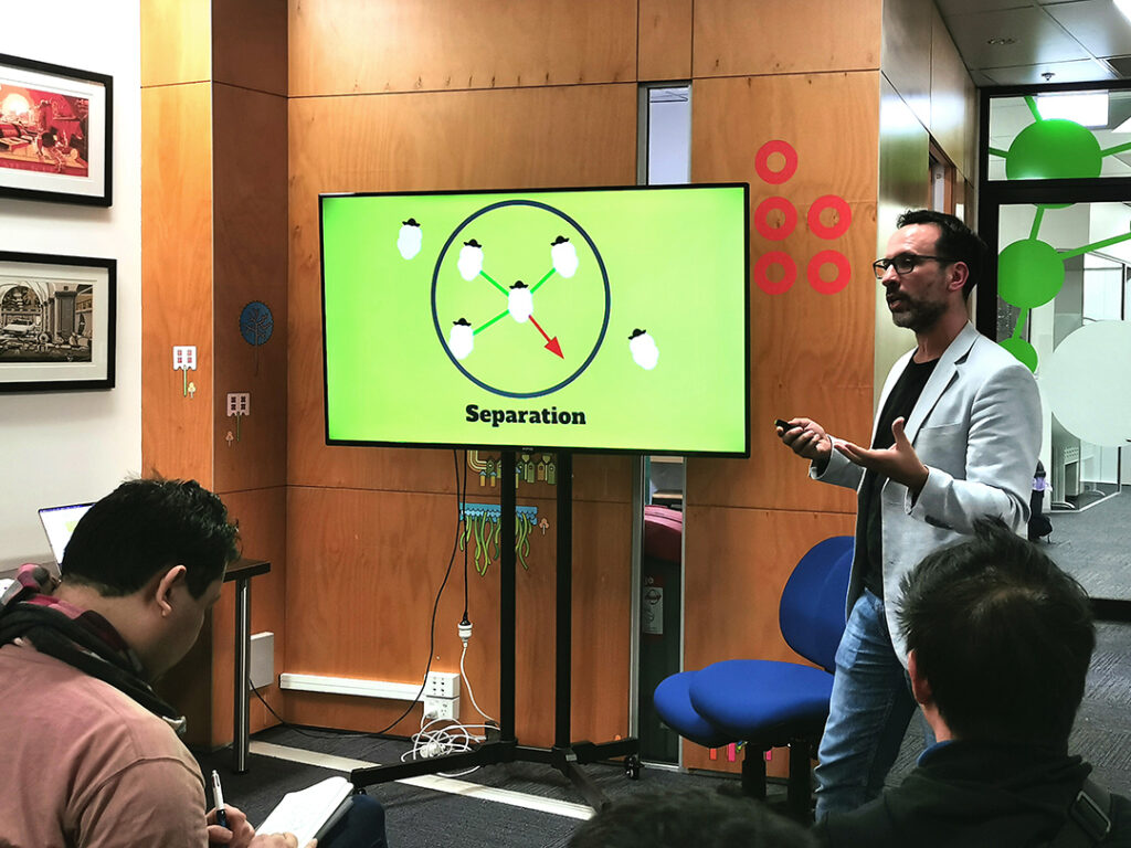 Another photo of David talking about the Boids principles in game. On screen is a slide with the "Separation" interaction, with an accompanying illustration of 4 sheep in a circle. 3 green lines connect 3 sheep to the one in the middle, but the one in the middle as a red arrow pointing to the empty space to the bottom left corner of the circle. David was using this to explain how sheep would be less inclined to leave this circle, despite the space available in it, as the herds moved in groups.